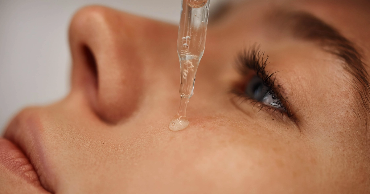 Glass dropper dispensing clear serum onto a person's skin near the blue eye for BBL Forever Young in O'Neill, NE. Focus is on the dropper and the liquid drop.