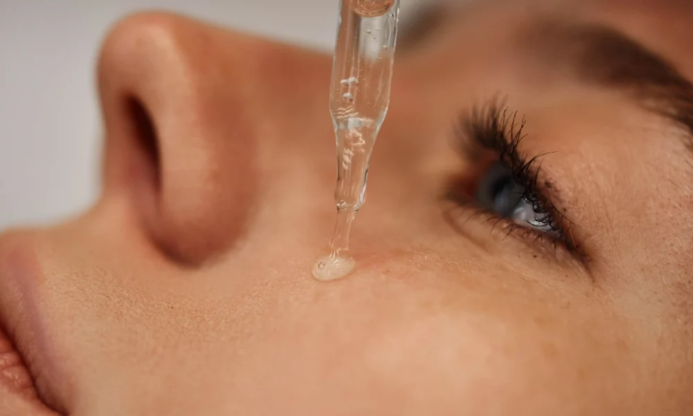 Glass dropper dispensing clear serum onto a person's skin near the blue eye for BBL Forever Young in O'Neill, NE. Focus is on the dropper and the liquid drop.